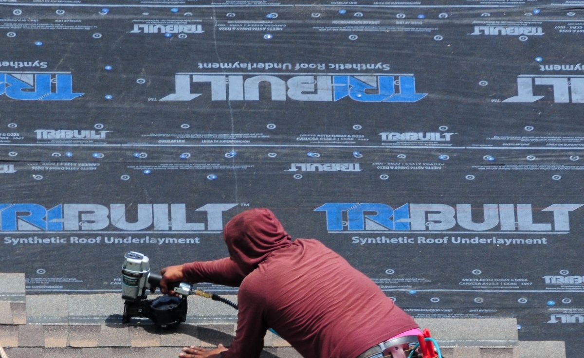 Roofer at work using a pneumatic nail gun to install shingles over a synthetic underlayment on a building’s roof